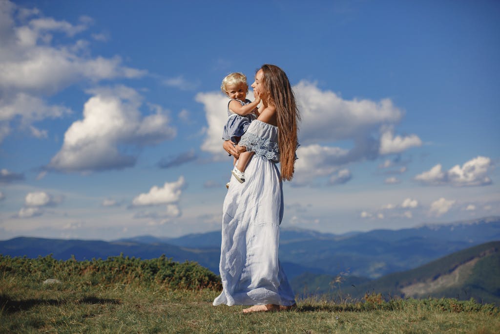 Mother and child in summer dress stand on a scenic hilltop under a blue sky.